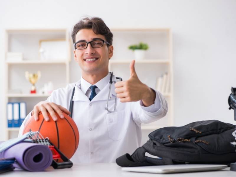 Médico sentado segurando uma bola de basquete fazendo sinal de positivo.