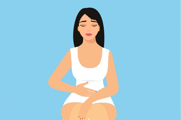 Na imagem, uma mulher vestida de branco com feição incomodada está com a mão na barriga.