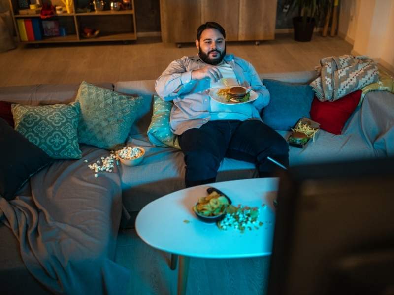 Homem obeso e sedentário sentado no sofá comendo fast-food.