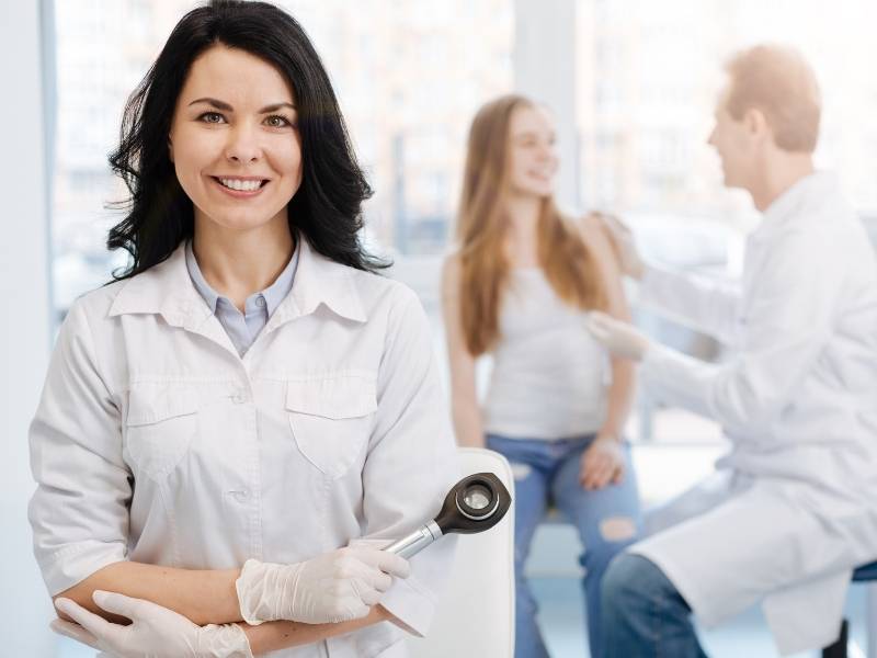 Mulher médica dermatologista com paciente ao fundo.