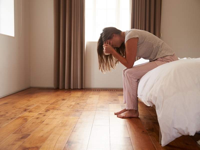 Mulher jovem sentada na cama e com as mãos no rosto.
