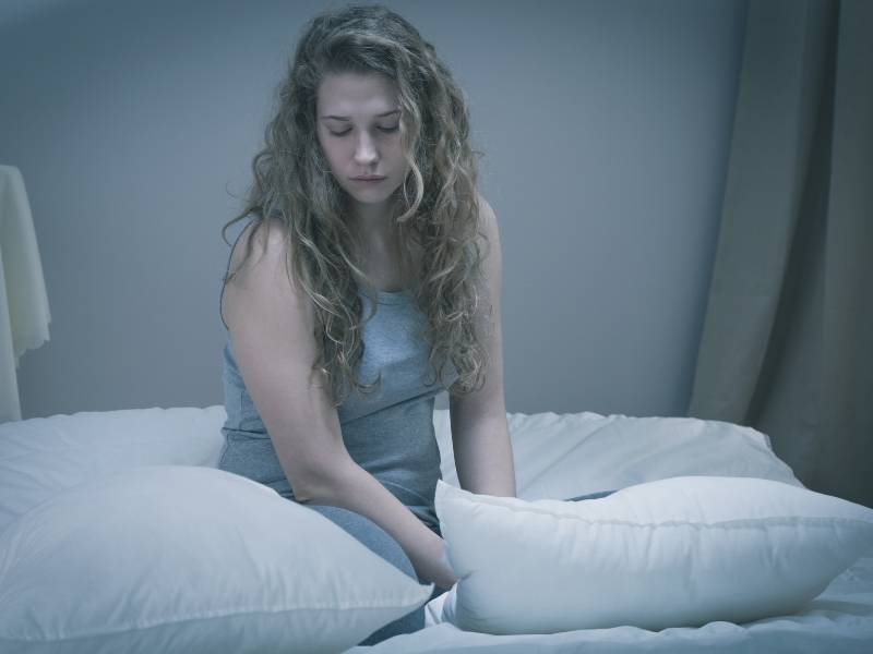 Mulher jovem sentada na cama com feição triste.