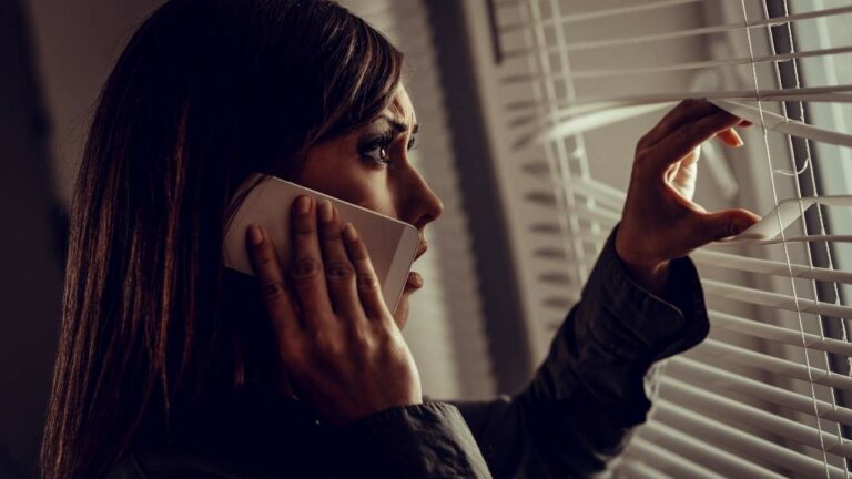 Mulher com medo olhando pela janela enquanto fala ao telefone.