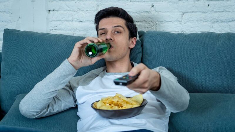 Homem sedentario sentado e bebendo no sofá.